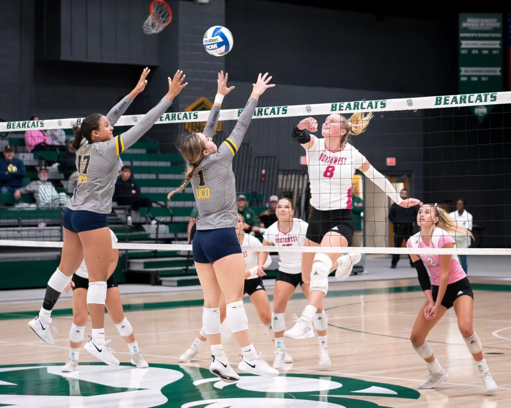 A cheerful college volleyball player hits the ball over the net toward opposing blockers.